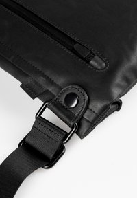 Black fabric bag featuring a smooth texture, a top zip closure, and a strap with metal hardware and a button closure detail.