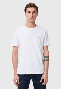 Valkoinen puuvillapaita, jossa on pyöreä kaula-aukko ja lyhyet hihat, rinnassa pieni musta logo. Yhdistetty tummiin farkkuihin.