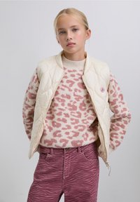 Cremefarben gesteppte Weste über einem rosa gepunkteten Pullover, kombiniert mit rosa Jeans im Zebramuster. Die Weste ist mit einem runden Logoknopf versehen.