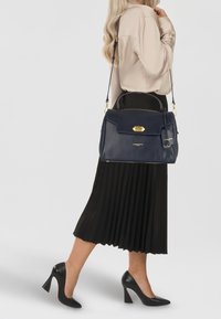 Borsa a tracolla in pelle blu navy con hardware dorato, forma strutturata, patta anteriore e tracolla removibile. Indossata con una gonna nera plissettata e tacchi.