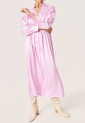 Osoba ubrana w jasnoróżową sukienkę midi z długim rękawem i guzikami, w połączeniu z beżowymi botkami na obcasie do połowy łydki, stojąca z założonymi skrzyżowanymi ramionami.