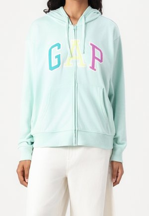 Person trägt eine hellblaue Kapuzenjacke mit Reißverschluss, auf der Brust mit mehrfarbigen "GAP"-Buchstaben, und weiße Hosen vor weißem Hintergrund.