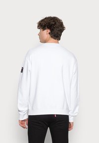 Witte sweatshirt met lange mouwen en een ronde halslijn. Heeft een zwart logopatch op de linkermouw en ribgebreide manchetten en zoom.