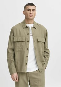 Chemise boutonnée vert olive en tissu texturé, dotée de deux poches poitrine et d'un col classique, portée par-dessus un t-shirt blanc.