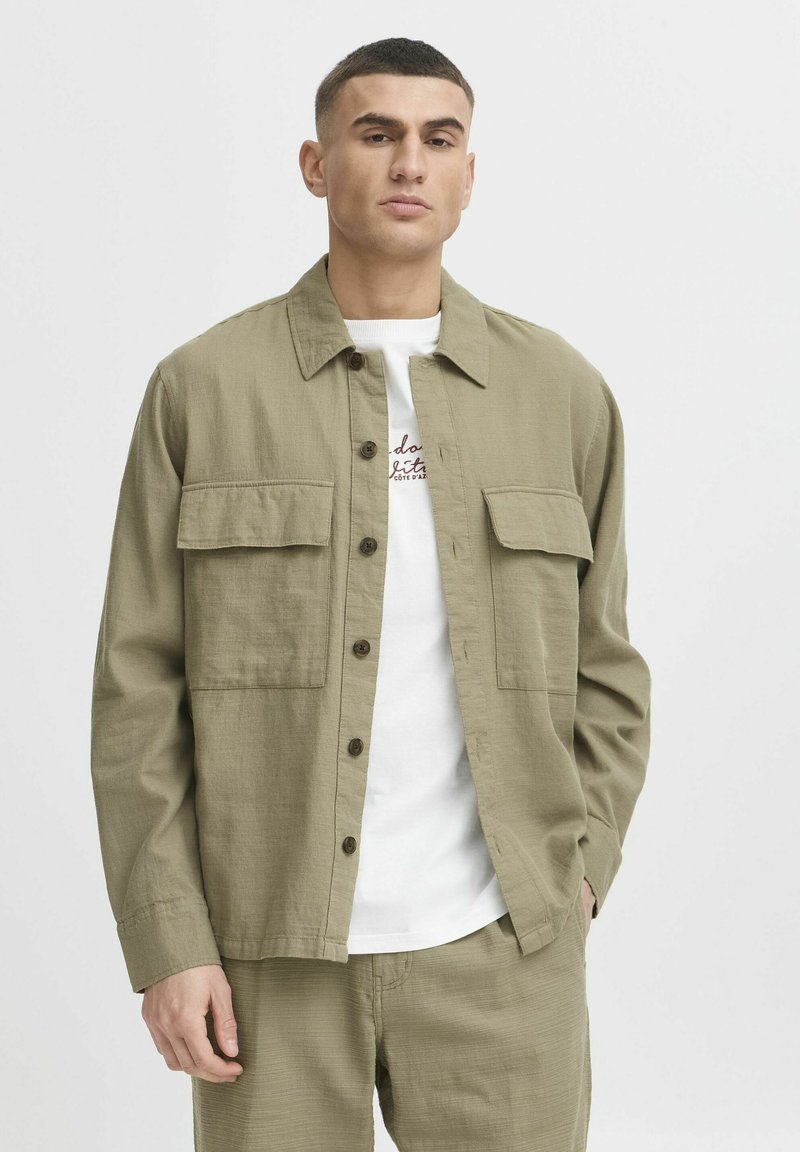 Chemise boutonnée vert olive en tissu texturé, dotée de deux poches poitrine et d'un col classique, portée par-dessus un t-shirt blanc.