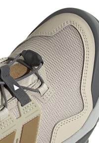 Primo piano della punta di una scarpa da trekking beige e marrone con tessuto in rete, cuciture rinforzate e chiusura a toggle su lacci grigi.