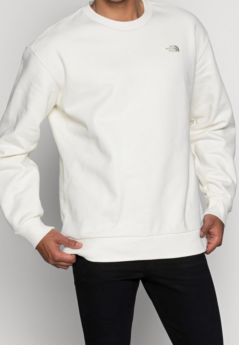Sweatshirt blanc en tissu doux, avec un col rond et des poignets côtelés, arborant un petit logo sur le haut du côté gauche de la poitrine.
