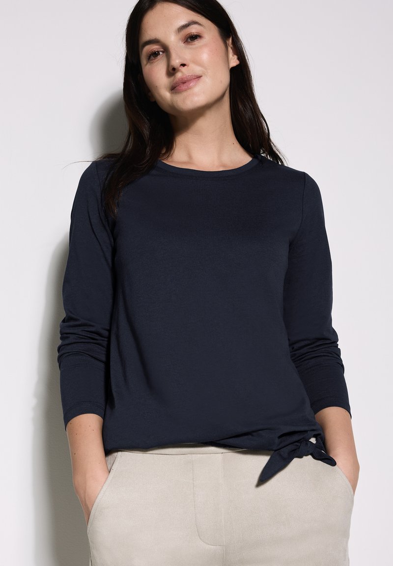 Maglietta a maniche lunghe blu navy con scollatura rotonda e dettaglio con nodo laterale arricciato, abbinata a pantaloni chiari e strutturati. Finitura in tessuto liscia.