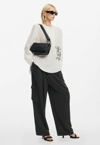Blouse blanche avec détail de boutons et broderie florale, assortie à un pantalon cargo noir ample. Les accessoires comprennent un sac bandoulière noir et des lunettes de soleil.