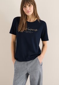 Femme en t-shirt bleu marine avec texte doré et pantalon gris à carreaux, debout les mains dans les poches devant un fond neutre.