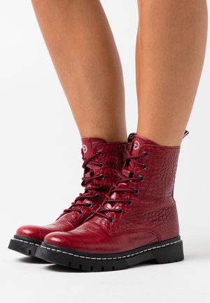 Bottes rouges à hauteur de cheville avec texture crocodile et lacets, semelles épaisses noires, portées sur des jambes nues sur un fond blanc.