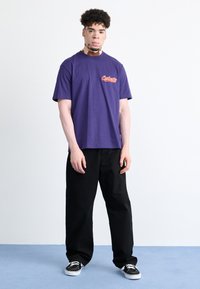 Purple cotton t-shirt with a front logo, short sleeves, and a crew neck paired with black wide-leg pants and black sneakers.