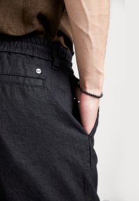 Pantalons en lin noirs avec une taille élastique, poches latérales et un petit logo circulaire, associés à une chemise marron.
