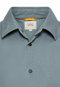 Męska koszula w jasnoniebieskim kolorze, z klasycznym kołnierzykiem, czarnymi guzikami i kieszenią na piersi. Wykonana z gładkiej, teksturowanej tkaniny.