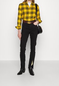 Donna con una camicia a quadri giallo e nero, pantaloni neri con fessure frontali che mostrano calzini a righe, stivali neri e che porta una borsa a tracolla nera.