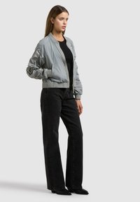 Jeune femme de profil portant une veste bomber gris clair, un haut noir, un jean noir taille haute à jambes larges, et des chaussures noires.