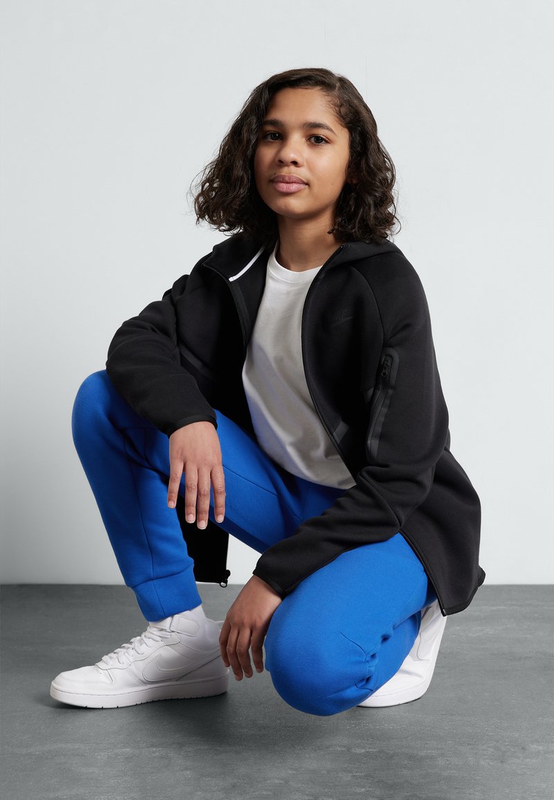 Schwarze Zip-Jacke mit hohem Kragen über einem weißen T-Shirt, kombiniert mit blauen Jogginghosen und weißen Sneakers, auf einer grauen Fläche posierend.