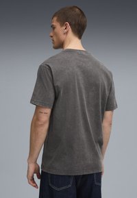 Szary t-shirt z krótkim rękawem o wyblakłej fakturze, z okrągłym dekoltem i widocznymi detalami szwów. Noszony z ciemnoniebieskimi dżinsami.