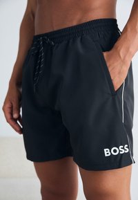 Black swim shorts made of lightweight material, featuring an elastic waistband with a drawstring, side pockets, and a white logo on the hem.