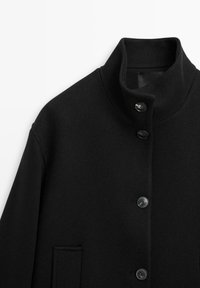 Black wool coat with a high collar, featuring five black buttons and a single patch pocket. The fabric has a textured finish.