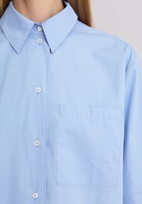 Jasnoniebieska koszula z guzikami, wykonana z gładkiej bawełny, z kołnierzykiem w kształcie spiczastym, pojedynczą kieszenią na piersi oraz białymi guzikami z przodu.