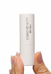 Witte cilindrische lipproduct met een gladde textuur, met gouden tekst "ZARKO BEAUTY BY OLI." Vastgehouden tussen vingers tegen een witte achtergrond.
