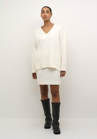 Maglione bianco lavorato a maglia con un motivo a diamante, scollatura a V e polsini a costine, abbinato a una gonne di maglia. Stivali neri fino al ginocchio con cinturini.