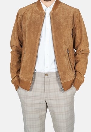 Homme portant une veste bombardier en suede beige par-dessus une chemise blanche à boutons et un pantalon à carreaux beige, les mains dans les poches.