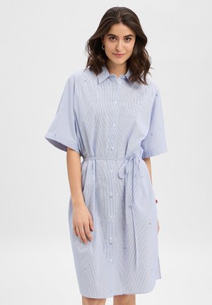 Młoda kobieta o brązowych, sięgających do ramion włosach, ubrana w jasnoniebieską sukienkę na guziki z krótkimi rękawami i wiązanym paskiem w talii.