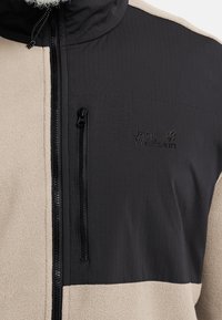 Fleecejack in zwart en beige. Voorzien van een hoge kraag, een borstzak met rits aan de voorkant, een gestructureerde stof en een geborduurd logo op de borst.