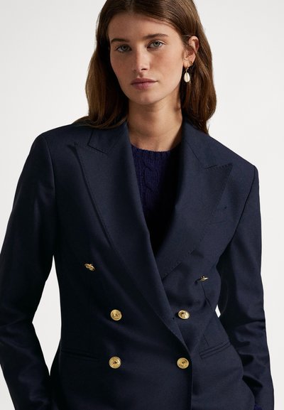 Młoda kobieta o brązowych włosach, ubrana w granatową, dwurzędową marynarkę z złotymi guzikami oraz ciemnoniebieski sweter z dzianiny pod spodem.