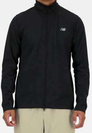 Man draagt een zwarte New Balance jas met ritssluiting en hoge kraag, met beige broek, staand tegen een effen achtergrond.