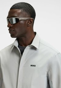Sunglasses with gray frames and dark mirrored lenses, featuring a logo. Worn with a light gray collared shirt with a subtle texture.