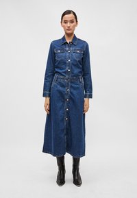 Denim skjortekjole i mørkeblå, med knappelukking foran, lange ermer, to brystlommer, og en avslappet passform med en lengde under kneet.