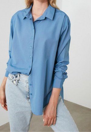 Kobieta ubrana w luźną, jasnoniebieską koszulę na guziki z podwiniętymi rękawami oraz jasne jeansy z detalem przewiązanego szlufki.