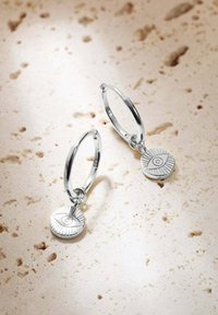 Orecchini a cerchio in argento con pendenti circolari caratterizzati da un motivo a raggi di sole e un occhio. Lo sfondo è una pietra chiara e screziata.