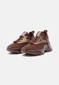 Un paio di sneaker a maglia marroni con suole spesse, lacci elastici a bungee e accenti bronzo metallico su uno sfondo bianco.