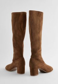 Bottes montantes en daim marron avec une texture lisse, un bout carré et un talon bloc robuste, caractérisées par des coutures minimalistes et une silhouette ajustée.