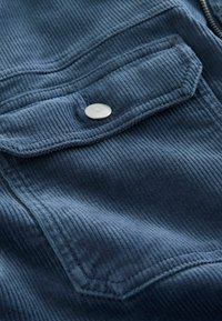 Poche d'une veste en velours côtelé bleu avec rabat et bouton-pression métallique, montrant le tissu texturé et une partie d'une fermeture éclair sur le côté droit.