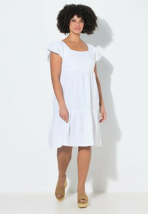 Kvinde med krøllet hår iført en hvid knælange kjole og beige kilehæle sandaler, stående mod en lys baggrund.
