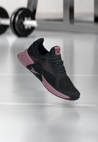 Μαύρο και ροζ αθλητικό παπούτσι Reebok που αιωρείται πάνω από γυαλιστερό πάτωμα με θολά βάρη γυμναστηρίου στο παρασκήνιο.