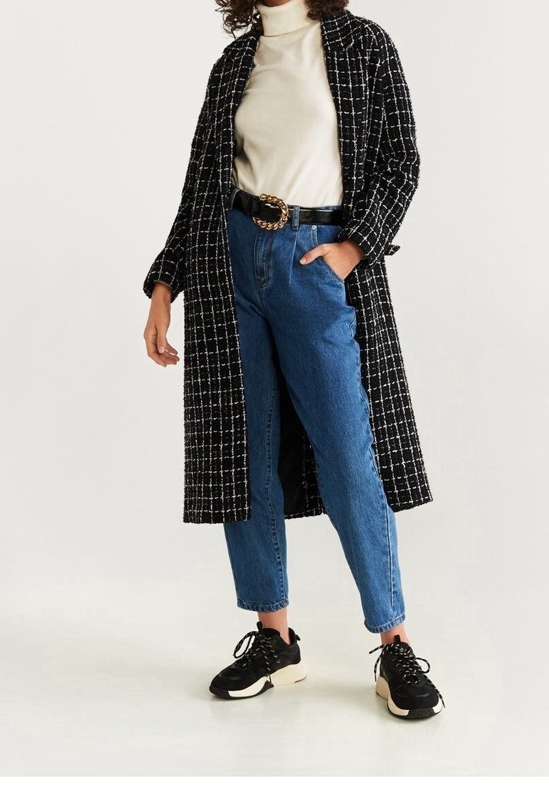 Manteau long à carreaux noir et blanc, col roulé crème, jean bleu taille haute, baskets noires avec semelles blanches, et une large ceinture avec une boucle dorée.