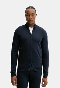 Uomo con i capelli intrecciati che indossa una giacca nera con zip sopra una camicia bianca, in piedi contro uno sfondo chiaro e semplice.