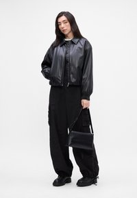 Giacca cropped in finta pelle nera con polsini elastici, abbinata a pantaloni cargo neri larghi e una piccola borsa a tracolla nera. Texture liscia.