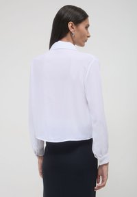 Donna con lunghi capelli scuri, che indossa orecchini a cerchio argentati, una camicia bianca a maniche lunghe e una gonna nera, mostrata di spalle.