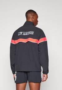Black athletic jacket with a high collar, featuring a pink horizontal stripe and reflective accents. Back displays "tcs LONDON MARATHON 2025" text.