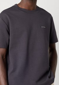 Czarnoszary bawełniany t-shirt z krótkim rękawem, okrągłym dekoltem i naszywką z logo Calvin Klein na lewej piersi. Gładka faktura, luźny krój.