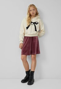 Cremefarbener Kapuzenpullover mit schwarzem Schleifen-Graphic, kombiniert mit einem bordeauxroten Samtrock, der goldene Punkte hat, und schwarzen Schnürstiefeln im combat Stil.