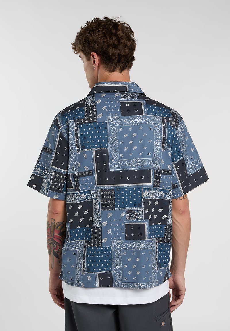 Camicia a maniche corte realizzata in tessuto con un design patchwork in diverse tonalità di blu e nero, con motivi e accenti bianchi.