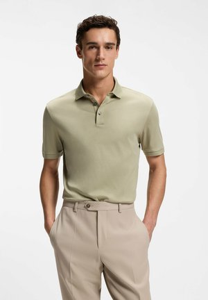 Junger Mann mit einem hellgrünen kurzärmeligen Poloshirt und beigen Hosen steht mit den Händen in den Taschen vor einem einfarbigen Hintergrund.